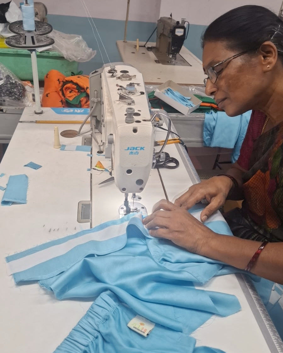 A seamstress at a Jack industrial machine in the Zouhi workshop outside Kochi, sewing the pale blue striped cloth of the Pale Sky Co-ord. The woven label is visible on the finished piece in the foreground.
