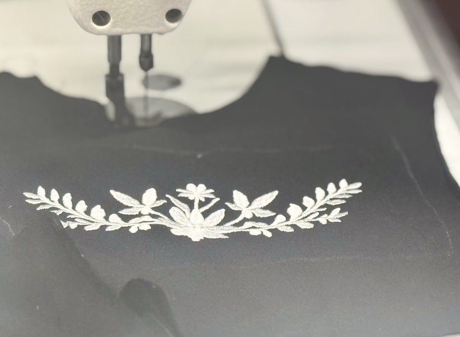 White floral embroidery worked into dark cloth, running beneath the needle of an embroidery machine.